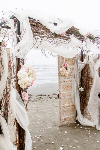 old door wedding decoration on the beach white doors with flowers decorations with wooden branches and cloth c baron photography via instagram