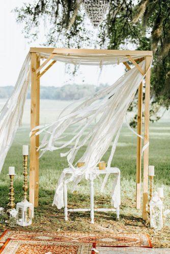 hippie wedding arch of light brown wood decorated with white ribbons carpet with ornament and candlesticks the portos