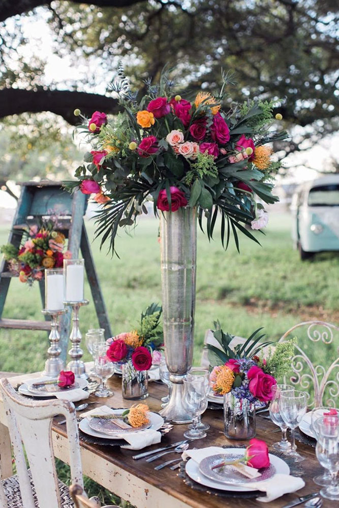 tall wedding centerpieces in a high metallic silvery vase bright burgundy and yellow roses with greens at a reception in boho style elisheva golani via instagram