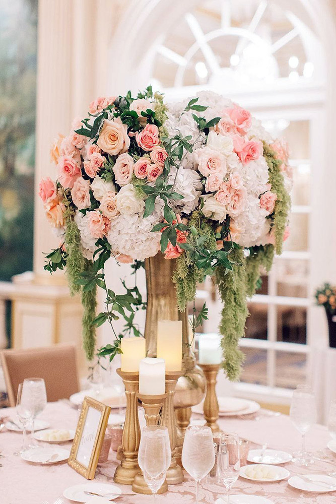 tall wedding centerpieces in a tall golden vase surrounded by candles a bouquet of coral roses and greens cascade kenzie victory via instagram
