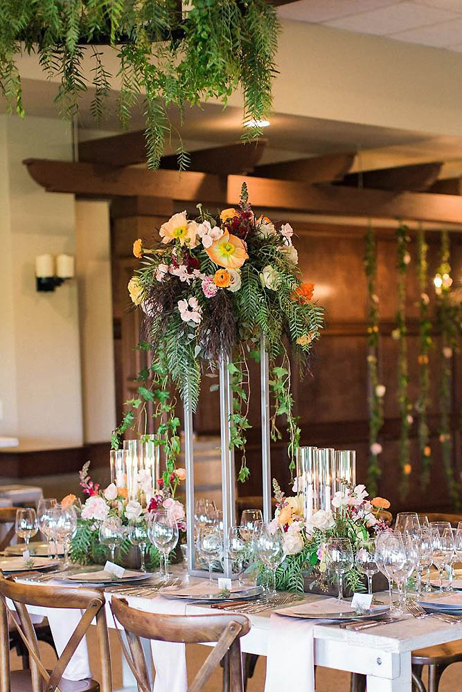tall wedding centerpieces on a wooden table with flowers and greens ashley bee via instagram