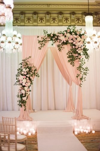 dusty rose wedding indoor ceremony with arch decorated with roses greenery and cloth candle aisle elisa bricker
